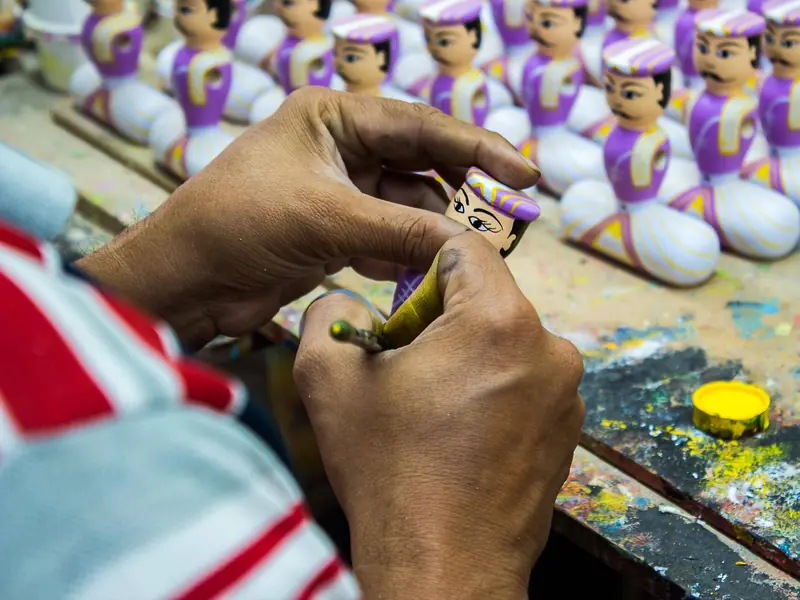 An artisan hand-painting a wooden doll, with rows of finished dolls behind, in Channapatna